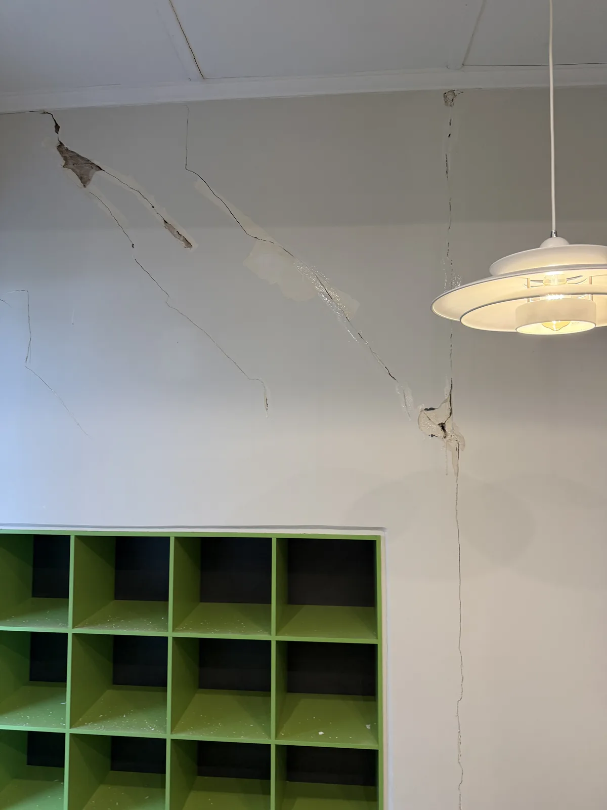 Interior wall above green shelving before repair, showing long visible cracks and damaged areas
